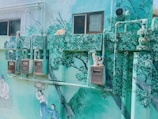 A mural on an outdoor wall depicts a serene natural scene, featuring painted trees and people dressed in traditional clothing. Real cats are sitting on top of the utility boxes mounted on the wall, which are painted to blend into the art.