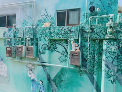 A mural on an outdoor wall depicts a serene natural scene, featuring painted trees and people dressed in traditional clothing. Real cats are sitting on top of the utility boxes mounted on the wall, which are painted to blend into the art.
