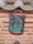A traditional clay roof tile with a detailed floral painting hangs on a textured wall. The tile features vibrant purple and blue flowers with green leaves against a dark background. The wall is made of a light terracotta-colored material with horizontal rows of protruding stone strips.