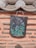 A traditional clay roof tile with a detailed floral painting hangs on a textured wall. The tile features vibrant purple and blue flowers with green leaves against a dark background. The wall is made of a light terracotta-colored material with horizontal rows of protruding stone strips.