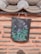 A traditional clay roof tile with a detailed floral painting hangs on a textured wall. The tile features vibrant purple and blue flowers with green leaves against a dark background. The wall is made of a light terracotta-colored material with horizontal rows of protruding stone strips.