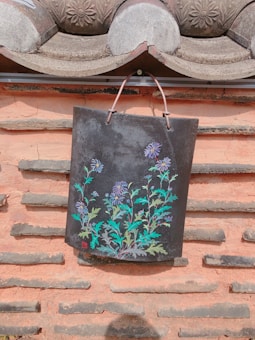 A traditional clay roof tile with a detailed floral painting hangs on a textured wall. The tile features vibrant purple and blue flowers with green leaves against a dark background. The wall is made of a light terracotta-colored material with horizontal rows of protruding stone strips.