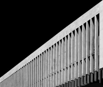 A modern architectural structure featuring a series of vertical concrete fins projecting from the facade against a stark black background. The repetitive pattern creates a striking visual effect with clean lines and a geometric design.