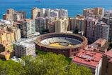 An urban landscape featuring a large circular bullring surrounded by high-rise residential buildings. The bullring has a clear view with an empty arena. In the background, a body of water, likely the sea, can be seen with a ship in the distance. Foreground foliage adds some greenery to the scene.