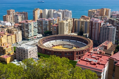 An urban landscape featuring a large circular bullring surrounded by high-rise residential buildings. The bullring has a clear view with an empty arena. In the background, a body of water, likely the sea, can be seen with a ship in the distance. Foreground foliage adds some greenery to the scene.