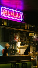 A dimly lit bar setting with a vibrant neon sign in Japanese characters hanging above the counter. Two staff members, one wearing a bucket hat and the other a headband, are working behind the bar. The decor includes various posters and bottles on the shelves, creating an eclectic and cozy atmosphere.