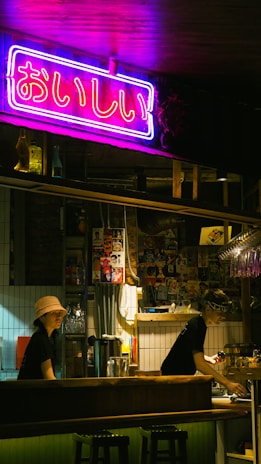 A dimly lit bar setting with a vibrant neon sign in Japanese characters hanging above the counter. Two staff members, one wearing a bucket hat and the other a headband, are working behind the bar. The decor includes various posters and bottles on the shelves, creating an eclectic and cozy atmosphere.