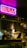 A dimly lit bar setting with a vibrant neon sign in Japanese characters hanging above the counter. Two staff members, one wearing a bucket hat and the other a headband, are working behind the bar. The decor includes various posters and bottles on the shelves, creating an eclectic and cozy atmosphere.