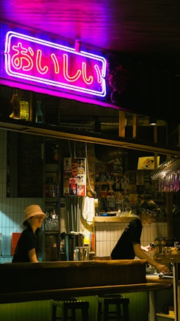 A dimly lit bar setting with a vibrant neon sign in Japanese characters hanging above the counter. Two staff members, one wearing a bucket hat and the other a headband, are working behind the bar. The decor includes various posters and bottles on the shelves, creating an eclectic and cozy atmosphere.