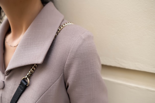 A close-up of a person wearing a textured beige or taupe jacket with a large collar. A gold and black chain strap is visible on the shoulder, suggesting a handbag or accessory.