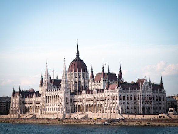 A grand, Gothic-style parliament building with intricate details, spires, and a large central dome situated by a riverbank. The structure is illuminated by natural daylight, highlighting its architectural features.