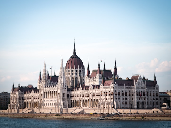 A grand, Gothic-style parliament building with intricate details, spires, and a large central dome situated by a riverbank. The structure is illuminated by natural daylight, highlighting its architectural features.