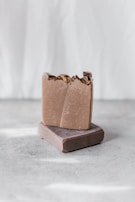 Two handmade soap bars are stacked on a textured gray surface. The top soap bar has a rustic, natural appearance with dried flowers embedded on the surface. The background is softly blurred in a light, neutral tone, giving a minimalist aesthetic.