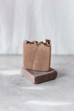 Two handmade soap bars are stacked on a textured gray surface. The top soap bar has a rustic, natural appearance with dried flowers embedded on the surface. The background is softly blurred in a light, neutral tone, giving a minimalist aesthetic.