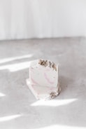 A stack of two artisanal soap bars with a creamy white base and subtle pink swirls. The soap is garnished with small dried herbs on the top. It is placed on a smooth surface with scattered light reflections.