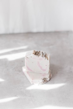 A stack of two artisanal soap bars with a creamy white base and subtle pink swirls. The soap is garnished with small dried herbs on the top. It is placed on a smooth surface with scattered light reflections.