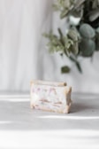 A bar of soap with scattered plant material and pinkish hues on a light surface. In the background, there are blurred green leaves adding a natural element to the scene.