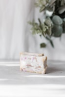 A bar of soap with scattered plant material and pinkish hues on a light surface. In the background, there are blurred green leaves adding a natural element to the scene.