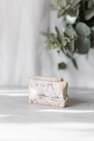 A bar of soap with scattered plant material and pinkish hues on a light surface. In the background, there are blurred green leaves adding a natural element to the scene.