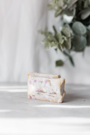 A bar of soap with scattered plant material and pinkish hues on a light surface. In the background, there are blurred green leaves adding a natural element to the scene.