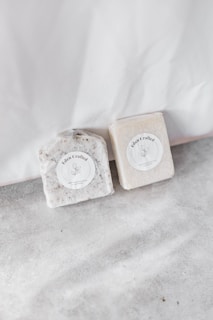 Two handcrafted soap bars are placed on a light, textured surface. Each bar has a round label with the words 'Eden Crafted' and a minimalist botanical illustration. The soaps appear to have natural, earthy tones and textures.