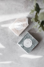Two artisan soap bars wrapped in translucent paper and labeled 'Eden Crafted' rest on a light, textured surface. One soap is a creamy white color, while the other has a deep green hue. Soft shadows and blurred leafy plants frame the soaps on the right side.