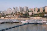 Cruise ships docked along a river with a backdrop of a densely populated cityscape featuring numerous high-rise buildings. Trees and greenery are interspersed among the buildings, and a concrete embankment separates the urban area from the water.