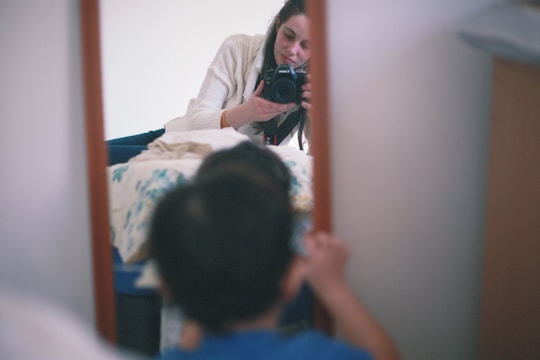 A person is taking a photograph using a DSLR camera, reflected in a mirror. In the foreground, a child is partially visible, looking towards the mirror. The setting appears to be indoors with a casual, home-like environment.
