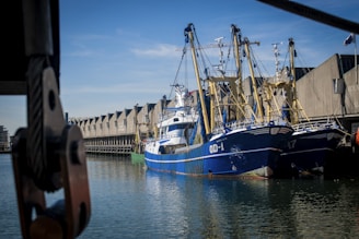 Two large fishing boats are docked at a harbor with a series of industrial buildings in the background. The boats are painted in shades of blue and white with some yellow accents. The calm water reflects the boats and buildings, while the sky is clear with a few clouds.