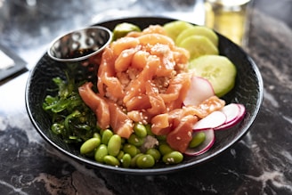 A black bowl contains a colorful poke dish featuring fresh, sliced salmon with sesame seeds, accompanied by edamame beans, sliced cucumbers, seaweed salad, and thin radish slices on a bed of rice. A metallic sauce cup is nestled inside the bowl. The setting is on a marble surface.