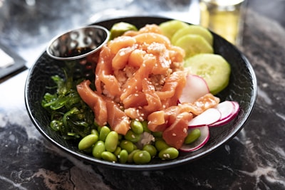 A black bowl contains a colorful poke dish featuring fresh, sliced salmon with sesame seeds, accompanied by edamame beans, sliced cucumbers, seaweed salad, and thin radish slices on a bed of rice. A metallic sauce cup is nestled inside the bowl. The setting is on a marble surface.