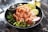 A black bowl contains a colorful poke dish featuring fresh, sliced salmon with sesame seeds, accompanied by edamame beans, sliced cucumbers, seaweed salad, and thin radish slices on a bed of rice. A metallic sauce cup is nestled inside the bowl. The setting is on a marble surface.