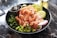 A black bowl contains a colorful poke dish featuring fresh, sliced salmon with sesame seeds, accompanied by edamame beans, sliced cucumbers, seaweed salad, and thin radish slices on a bed of rice. A metallic sauce cup is nestled inside the bowl. The setting is on a marble surface.