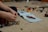 Hands of a person assembling a Lego construction with an instruction manual visible on a carpeted floor. Various colored Lego pieces are scattered around, including wheels and small brick structures.