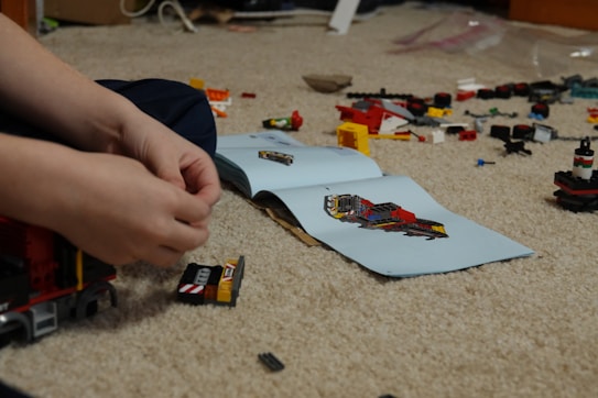 Hands of a person assembling a Lego construction with an instruction manual visible on a carpeted floor. Various colored Lego pieces are scattered around, including wheels and small brick structures.