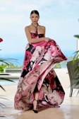 A model wearing an elegant, strapless gown with a vibrant floral pattern in shades of pink, black, and white. She stands poised with a serene expression against a blurred outdoor backdrop featuring greenery and a pool. Her hair is neatly styled, and she accessorizes with a beaded necklace and earrings.