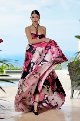 A model wearing an elegant, strapless gown with a vibrant floral pattern in shades of pink, black, and white. She stands poised with a serene expression against a blurred outdoor backdrop featuring greenery and a pool. Her hair is neatly styled, and she accessorizes with a beaded necklace and earrings.