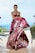 A model wearing an elegant, strapless gown with a vibrant floral pattern in shades of pink, black, and white. She stands poised with a serene expression against a blurred outdoor backdrop featuring greenery and a pool. Her hair is neatly styled, and she accessorizes with a beaded necklace and earrings.