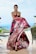A model wearing an elegant, strapless gown with a vibrant floral pattern in shades of pink, black, and white. She stands poised with a serene expression against a blurred outdoor backdrop featuring greenery and a pool. Her hair is neatly styled, and she accessorizes with a beaded necklace and earrings.