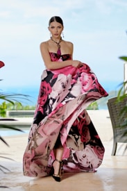 A model wearing an elegant, strapless gown with a vibrant floral pattern in shades of pink, black, and white. She stands poised with a serene expression against a blurred outdoor backdrop featuring greenery and a pool. Her hair is neatly styled, and she accessorizes with a beaded necklace and earrings.