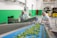 Two workers dressed in protective clothing are inspecting and sorting green leafy vegetables on a blue conveyor belt inside a factory setting. The environment is clean with green and white walls, and there are pieces of industrial equipment and yellow forklifts in the background.