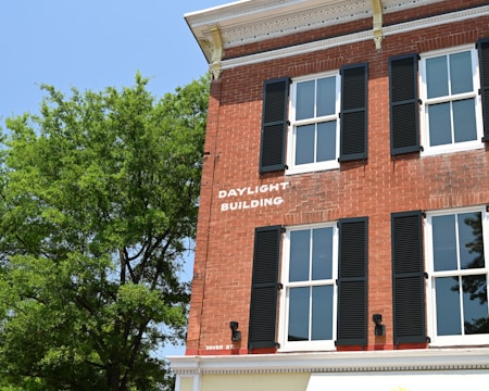 A red brick building with black shutters on the windows, labeled as 'DAYLIGHT BUILDING'. The structure is adjacent to a large green tree under a clear blue sky. Architectural details include decorative trim along the roofline.
