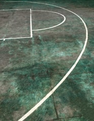 A basketball court featuring a white painted line curving on a concrete surface with a slightly worn and weathered appearance. The court's surface has a mix of green and gray hues, giving it a rustic look.