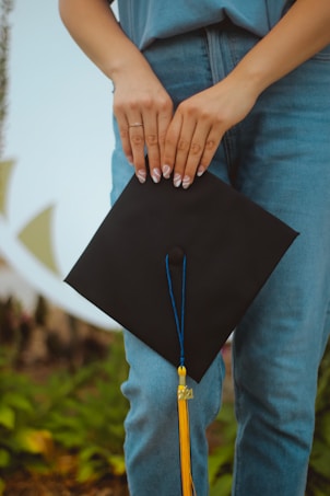 A person holding a black graduation cap with a blue tassel featuring a gold charm. The individual is wearing denim jeans and a blue shirt. The background shows blurred vegetation with green and yellow hues.