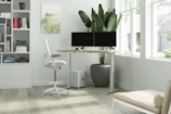 A modern, minimalist office space featuring a standing desk with dual monitors, a comfortable ergonomic chair, and a large potted plant beside the desk. Natural light streams in through large windows, illuminating a tidy bookshelf filled with various books. The room is decorated in light, neutral tones with a wooden floor and a simple, abstract piece of art on the wall.