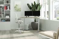 A modern, minimalist office space featuring a standing desk with dual monitors, a comfortable ergonomic chair, and a large potted plant beside the desk. Natural light streams in through large windows, illuminating a tidy bookshelf filled with various books. The room is decorated in light, neutral tones with a wooden floor and a simple, abstract piece of art on the wall.