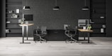 A modern office space with two workstations featuring adjustable desks and ergonomic chairs. The background includes a dark brick wall with symmetrical black shelves, adorned with minimalistic decor such as books, framed art, and plants. The lighting is provided by sleek pendant lamps.