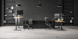A modern office space with two workstations featuring adjustable desks and ergonomic chairs. The background includes a dark brick wall with symmetrical black shelves, adorned with minimalistic decor such as books, framed art, and plants. The lighting is provided by sleek pendant lamps.