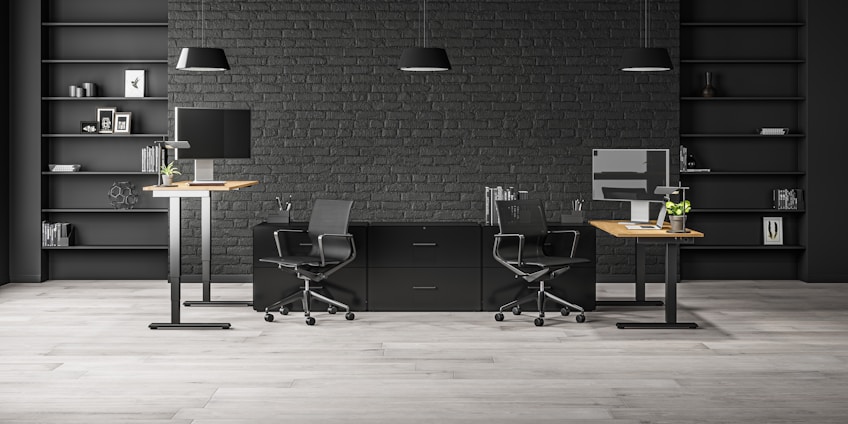 A modern office space with two workstations featuring adjustable desks and ergonomic chairs. The background includes a dark brick wall with symmetrical black shelves, adorned with minimalistic decor such as books, framed art, and plants. The lighting is provided by sleek pendant lamps.