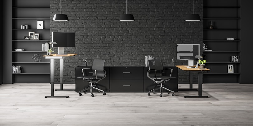 A modern office space with two workstations featuring adjustable desks and ergonomic chairs. The background includes a dark brick wall with symmetrical black shelves, adorned with minimalistic decor such as books, framed art, and plants. The lighting is provided by sleek pendant lamps.